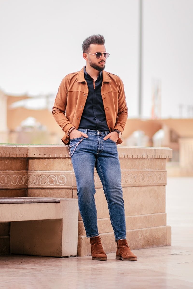 man in brown jacket and blue denim jeans sitting on brown concrete bench during daytime